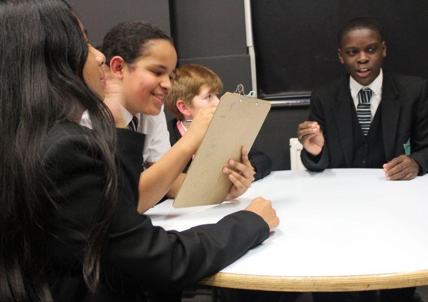 4 pupils in school uniform gathered around a table, one holding a clipboard, smiling