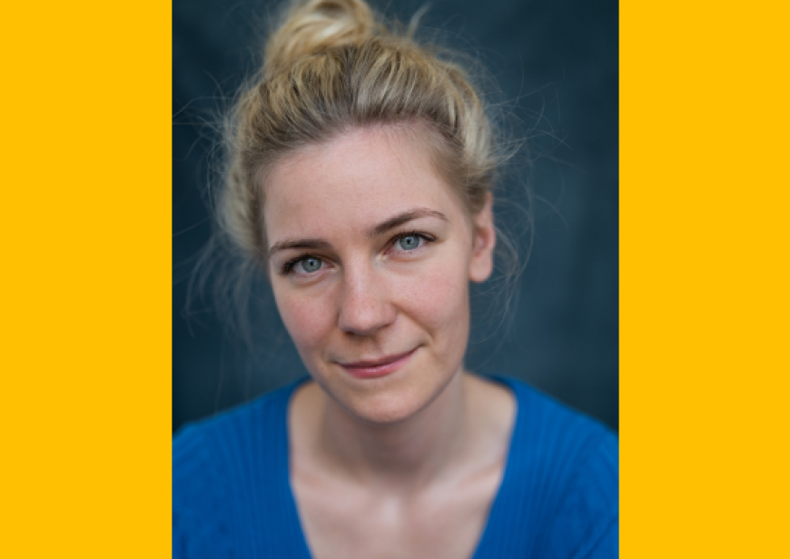 A headshot of a white woman of average height with long blonde hair pulled up in a messy bun wearing a blue v-neck top.