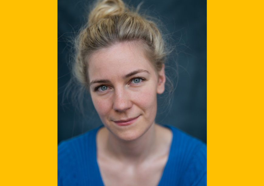 A headshot of a white woman of average height with long blonde hair pulled up in a messy bun wearing a blue v-neck top.  