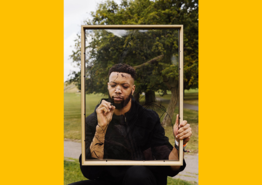 A person with short dark hair and a beard wearing a black top stands holding a frame in front of their face.