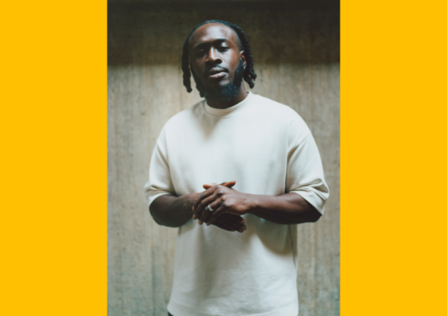 A person with dreads wearing a white t-shirt stands with hands clasped.