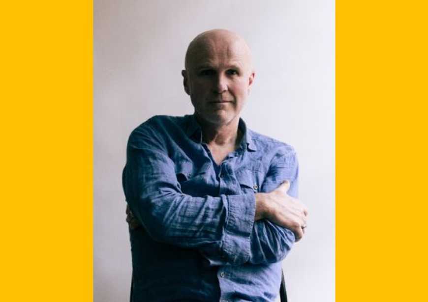 A white bald man sits wearing a blue long sleeved shirt with his arms crossed