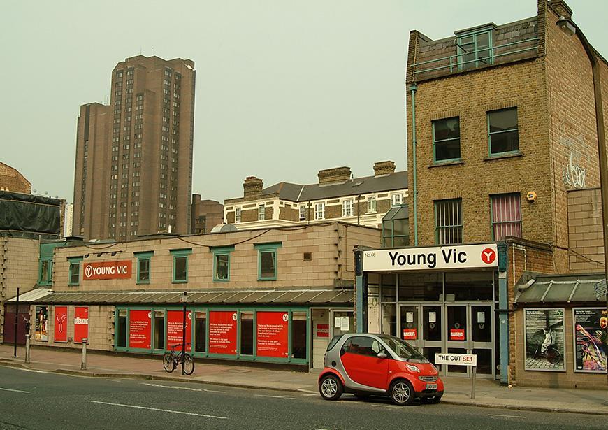 Exterior of the Young Vic. On the left side of the building is row of red posters. Outside the doors is The Cut roadside, and a red Smart car is parked alongside it.