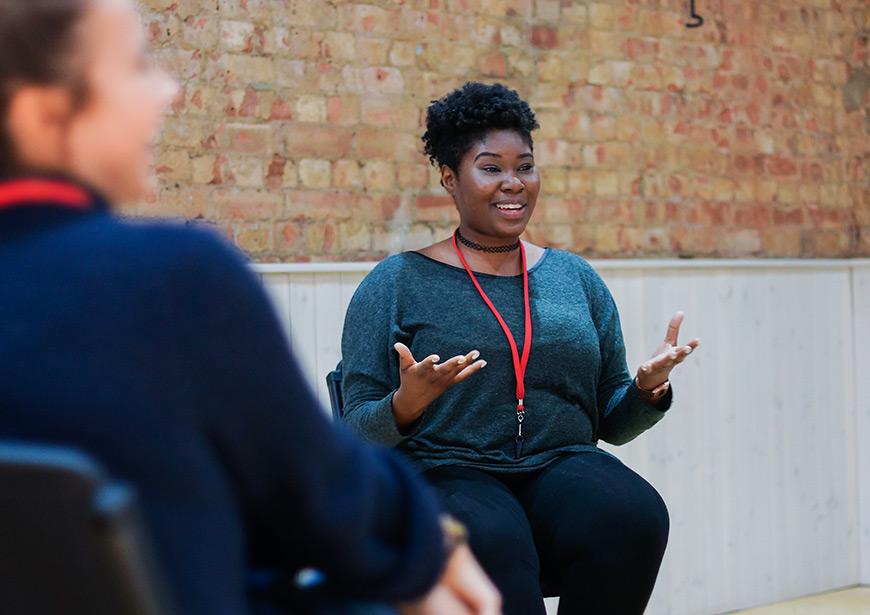 Workshop participant smiles with arms outstretched as they explain something to the group. Person in the foreground can be seen launching along.