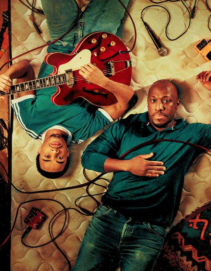 Viewed from above, two people lie side by side on an old mattress. The person on the left-hand side is lying upside down so their head is at the bottom of the image. They are wearing a green t-shirt with white stripes and hold a red electric guitar. The person on the right is lying the right way up, wearing a green collared long-sleeved shirt and has a red cable lying across their chest. The mattress is surrounded by cables, microphones, and other music equipment.