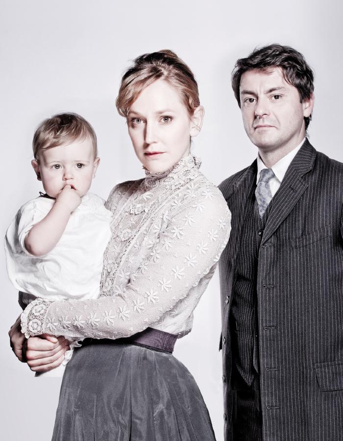 A woman and man stand together holding a baby, staring into the camera. They are all wearing period dress and look serious. The colours in the photo are muted, and the image had a pink boarder at the top and bottom. 