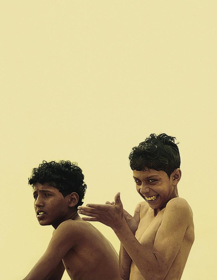 Two boys in a photograph. The boy to the left is looking into the distance with a slightly worried expression, the younger boy to the right is looking in a similar direction whilst rubbing his hands with a cheeky grin.