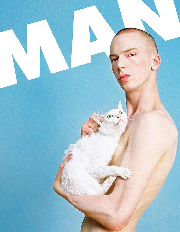 A naked man staring at the camera holding a white cat to his chest, against a baby blue background with the word 'MAN' emblazoned behind him in white 