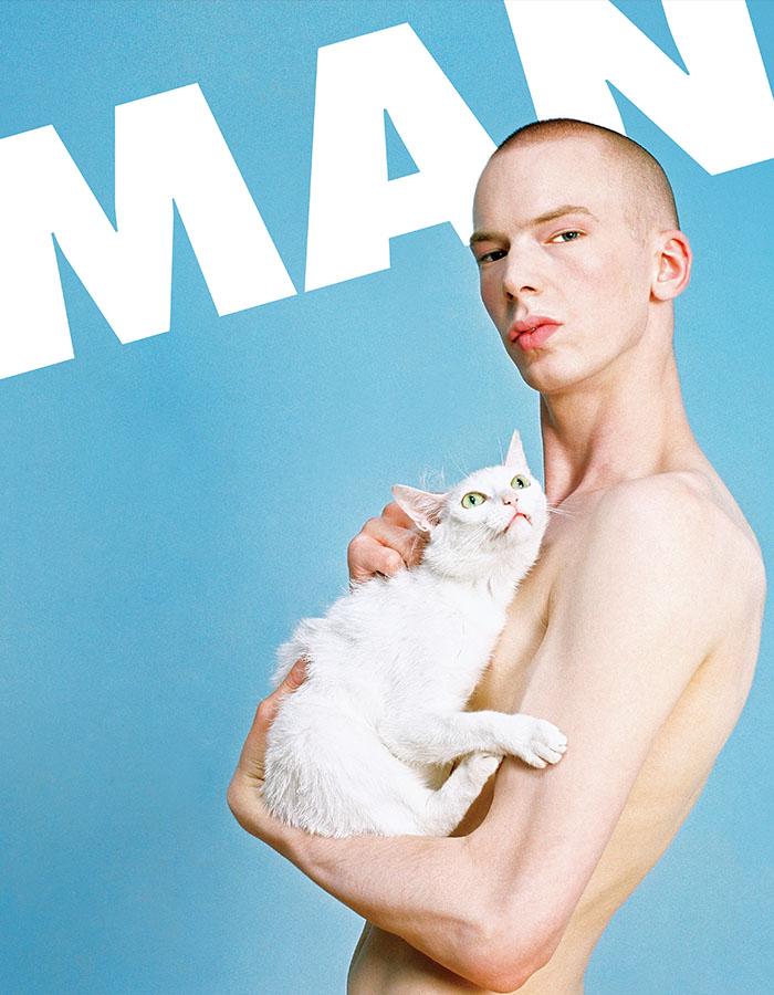 A naked man staring at the camera holding a white cat to his chest, against a baby blue background with the word 'MAN' emblazoned behind him in white