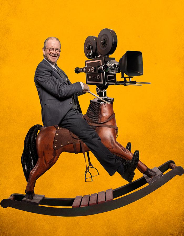 Harry Enfield riding a rocking horse with a old school film camera for a head