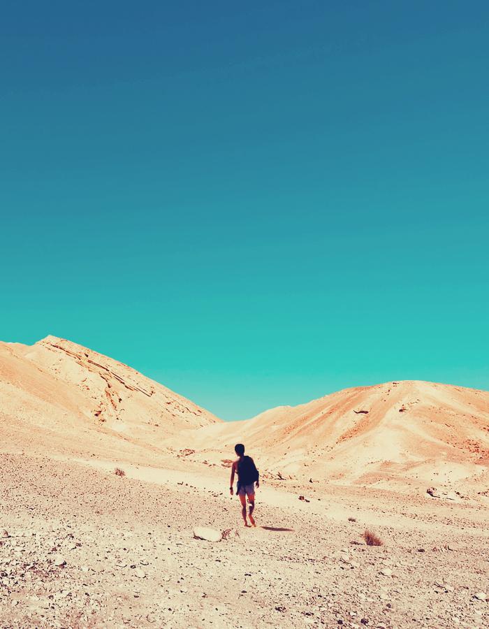 Boy walks through a desert towards two arid hills under a bright blue sky