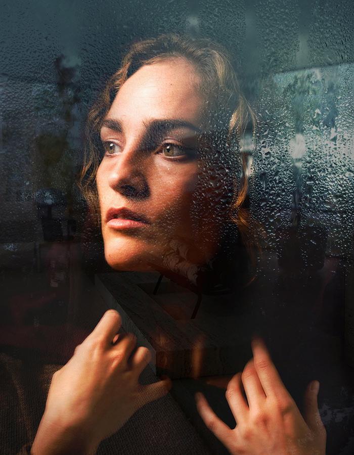 Woman looks pensively out window covered in rain drops 