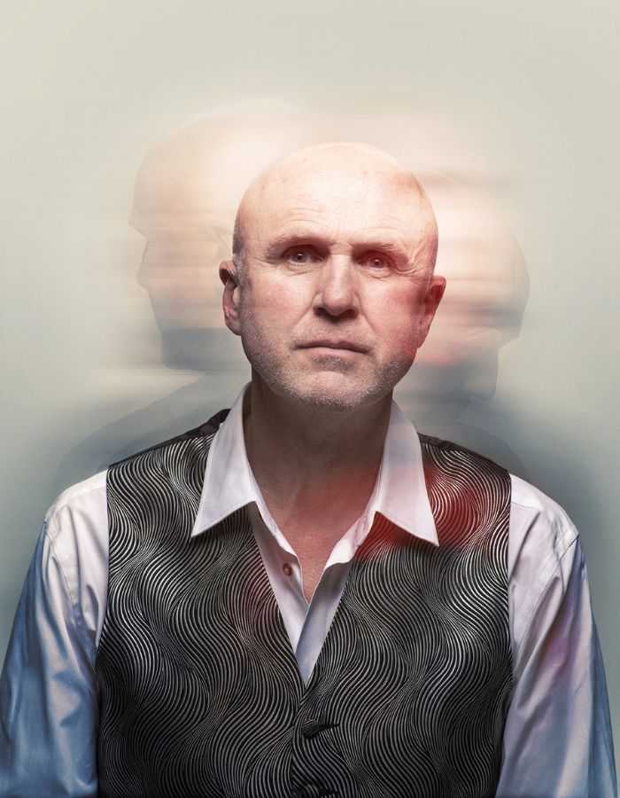 A head and shoulders shot of Tim Crouch wearing a white long sleeve shirt and a patterned gray vest. Bahind him are blurry shadows of his head facing the left and right.