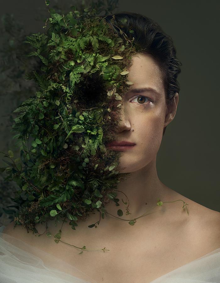 A sad looking bride played by Aoife Duffin looking straight at the camera.  She has a single teardrop about to fall from her right eye. The left side of her face is being overtaken with foliage. Photo by Sebastian Nevols. Concept by Emilie Chen. 