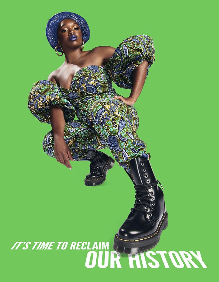 A woman wearing an intricately patterned jumpsuit, Doc Martin boots and a gele with her left hand on her hip crouches in front of a green background.