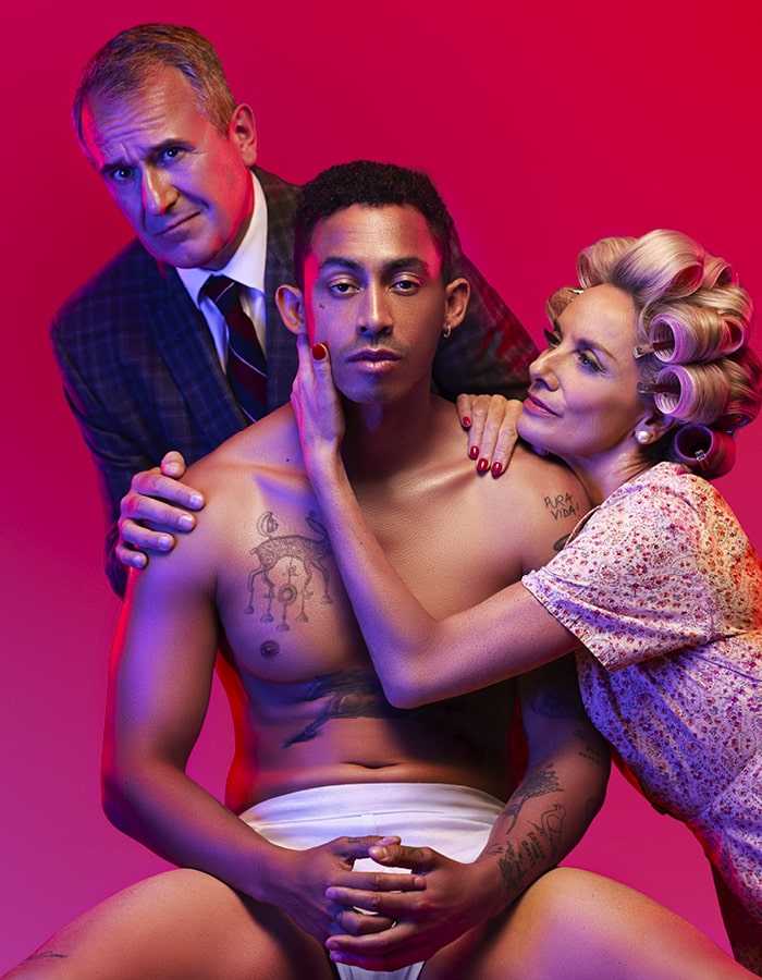 3 people pose together against a pink background. The person in the centre is sat on a chair wearing white underwear. A person kneels on the right with one hand cupping the face of the person in the middle. A person stands on left with both hands on the shoulder of the person in the centre.