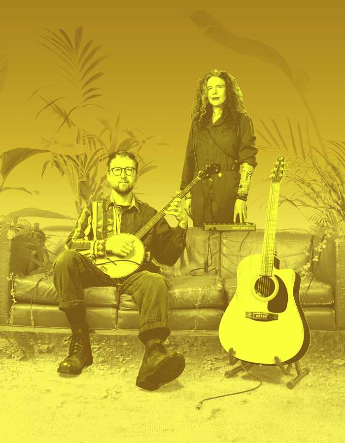 Against a yellow background are a man and woman. The man sits on a sofa and is holding a banjo; the woman stands behind the sofa. Next to them is a guitar on a stand.