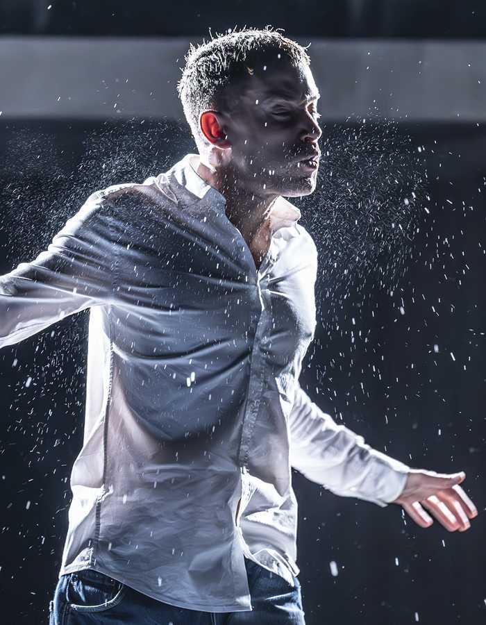 A man lit from behind, wearing a white shirt and blue jeans is standing on a stage. His arms are spread out to the side, and his eyes are closed. He is mid-action and there are sprays of water emanating from him.