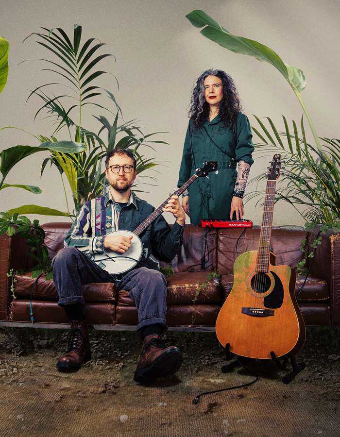 Against a background of green plants are a man and woman. The man sits on a sofa and is holding a banjo; the woman stands behind the sofa. Next to them is an orange guitar on a stand. 