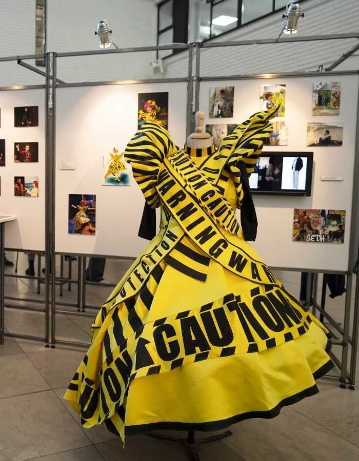 A picture of an exhibition with multiple photos on the walls and a dress on a mannequin made of black and yellow caution tape
