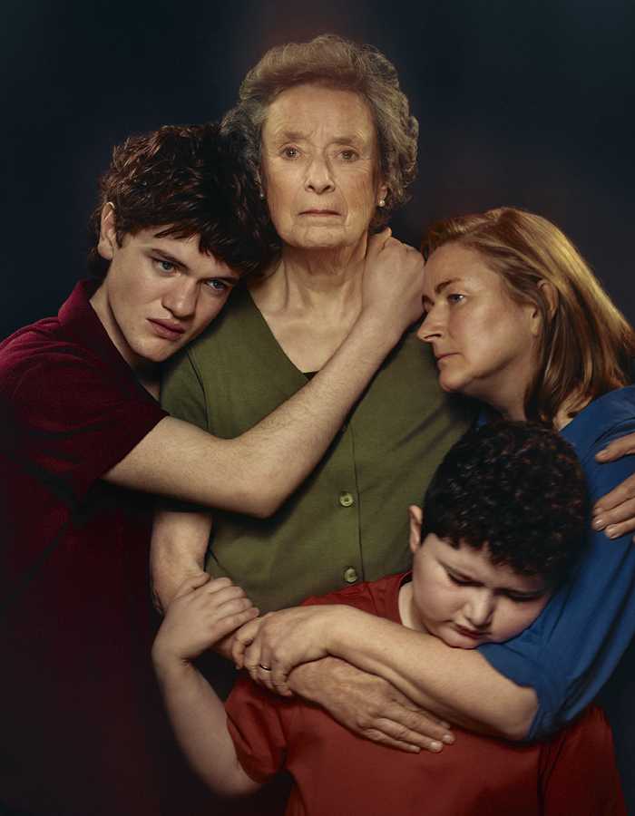 A family hug each other, Grandma in the centre, three family members surrounding her. She looks at the camera, everyone else is looking in different directions. They wear green, red, blue and marroon tops against a black backdrop