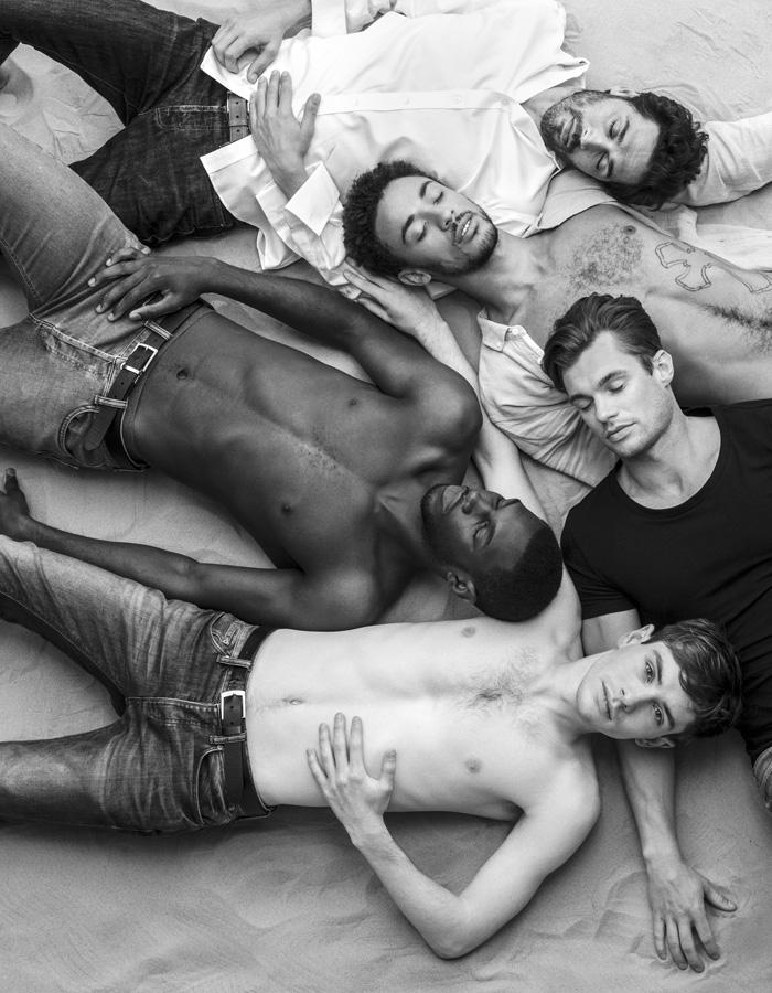 A group of 5 men lay on sand in various states on undress. One has his eyes open whilst the others have theirs closed.