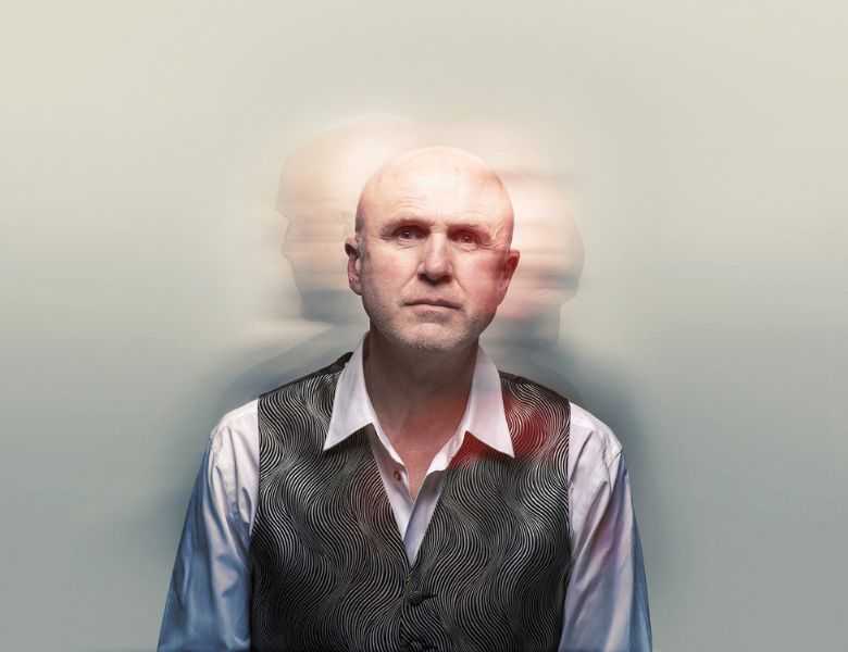 A head and shoulders shot of Tim Crouch wearing a white long sleeve shirt and a patterned gray vest. Bahind him are blurry shadows of his head facing the left and right. 