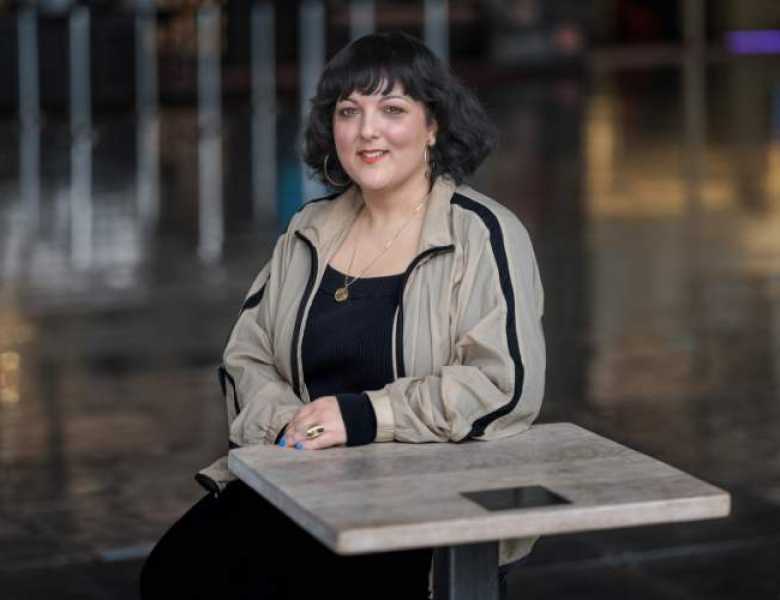 A light skinned, British South Asian woman with dark hair and a fringe, wearing a black top, beige jacket and gold jewellery, sat at a table