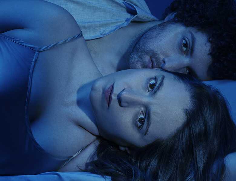 A man and a woman lie on a bed with light sheets. They both stare straight into the camera, awash in blue light. over the image, there is white text reading: Arthur Miller/Broken Glass/directed by Jordan Fein