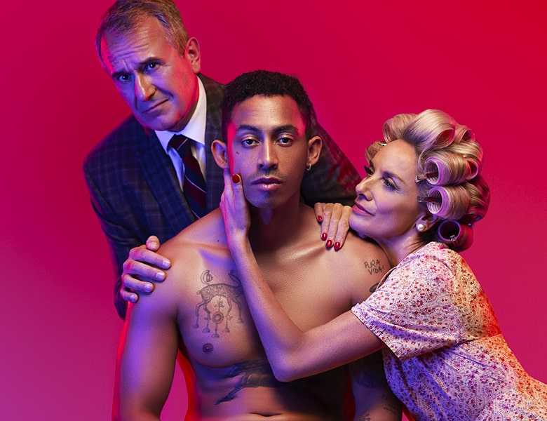 3 people pose together against a pink background. The person in the centre is sat on a chair wearing white underwear. A person kneels on the right with one hand cupping the face of the person in the middle. A person stands on left with both hands on the shoulder of the person in the centre.