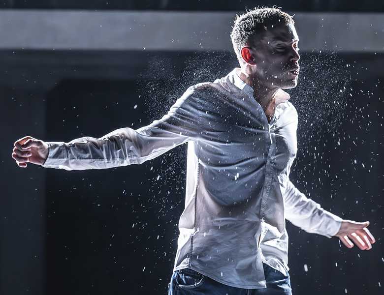A man lit from behind, wearing a white shirt and blue jeans is standing on a stage. His arms are spread out to the side, and his eyes are closed. He is mid-action and there are sprays of water emanating from him. 