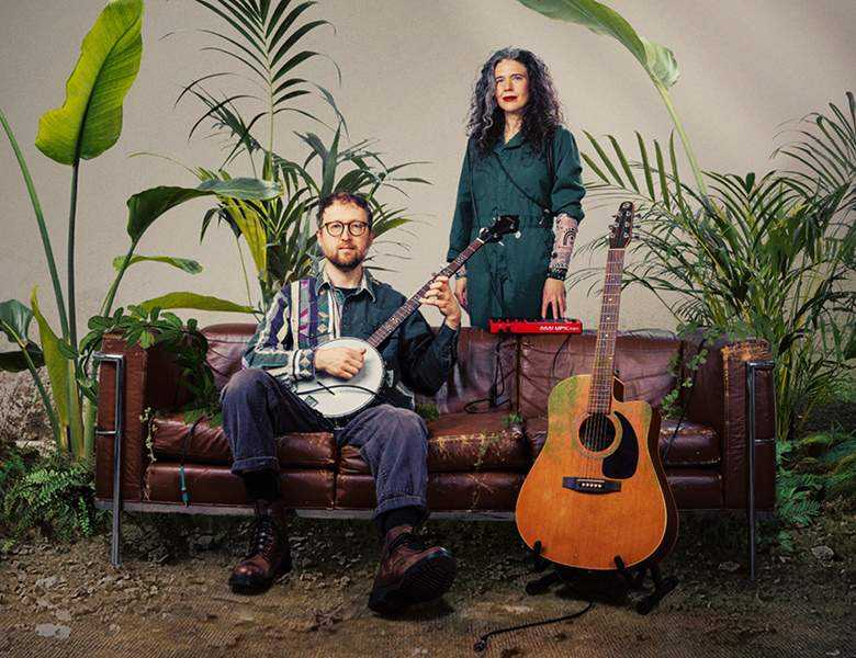 Against a background of green plants are a man and woman. The man sits on a sofa and is holding a banjo; the woman stands behind the sofa. Next to them is an orange guitar on a stand. 