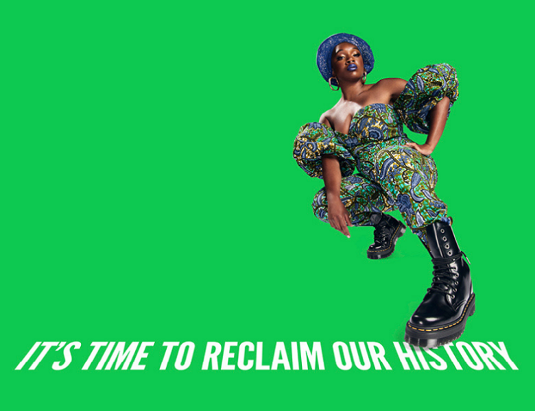 A woman wearing an intricately patterned jumpsuit, Doc Martin boots and a gele with her left hand on her hip crouches in front of a green background. Under her left foot are the words "it's time to reclaim our history". 
