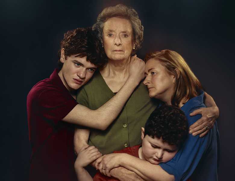 A family hug each other, Grandma in the centre, three family members surrounding her. She looks at the camera, everyone else is looking in different directions. They wear green, red, blue and marroon tops against a black backdrop