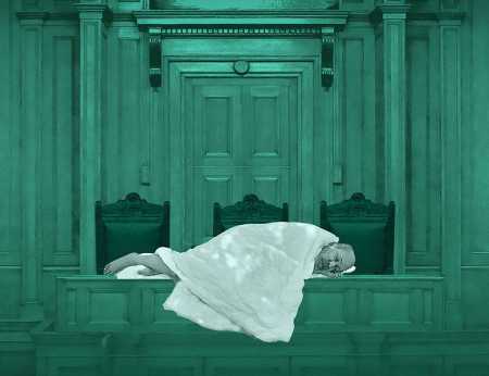 Against a teal background, an elderly man lies on a table in the House of Commons wrapped in a white blanket. 