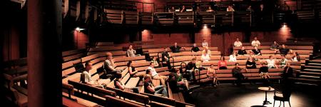 Main auditorium full of people, with an empty chair and desk in the middle. Photo by Philip Vile 