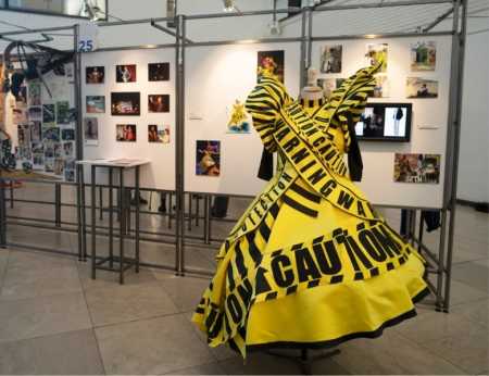 A picture of an exhibition with multiple photos on the walls and a dress on a mannequin made of black and yellow caution tape