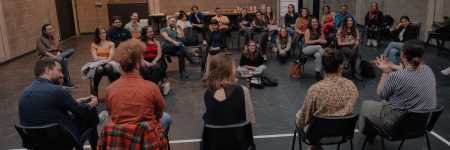A panel of five artists, with their backs turned to the camera, is speaking to an audience of about 30 people sat across from them.