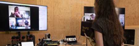 A workshop participant with long brown hair, wearing a black shirt and beige trousers interacting with remote participants on Zoom using a cube mic. In the background there is a large screenshowing Zoom details, and a table with remote conferencing equipment as part of the Tech Tower.