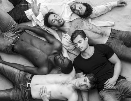 A group of 5 men lay on sand in various states on undress. One has his eyes open whilst the others have theirs closed.