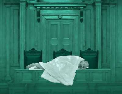 Against a teal background, an elderly man lies on a table in the House of Commons wrapped in a white blanket. 