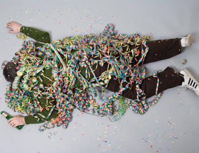 Man lies on a grey floor with beer bottle in hand, covered in confetti streamers