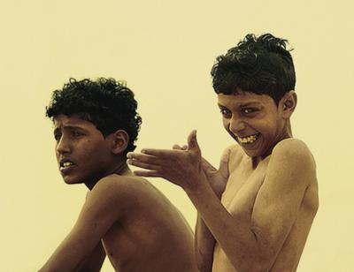 Two boys in a photograph. The boy to the left is looking into the distance with a slightly worried expression, the younger boy to the right is looking in a similar direction whilst rubbing his hands with a cheeky grin.