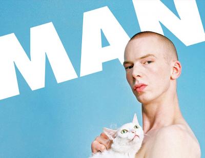 A naked man staring at the camera holding a white cat to his chest, against a baby blue background with the word 'MAN' emblazoned behind him in white