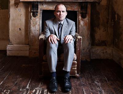 Rory Kinnear sits in a chair under a mirror in a derelict room 