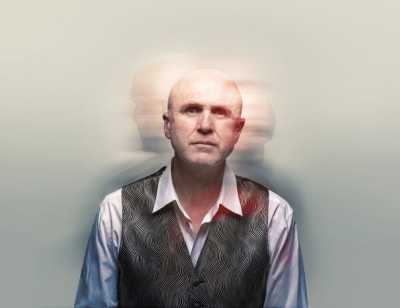 A head and shoulders shot of Tim Crouch wearing a white long sleeve shirt and a patterned gray vest. Bahind him are blurry shadows of his head facing the left and right. 
