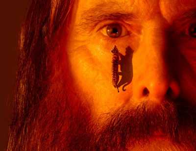 A close crop of a person's face with a full ginger beard and moustache lit with orange and red hues. The person has a tiger walking up their cheek towards their eye. 
