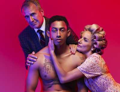 3 people pose together against a pink background. The person in the centre is sat on a chair wearing white underwear. A person kneels on the right with one hand cupping the face of the person in the middle. A person stands on left with both hands on the shoulder of the person in the centre.
