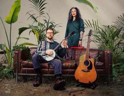 Against a background of green plants are a man and woman. The man sits on a sofa and is holding a banjo; the woman stands behind the sofa. Next to them is an orange guitar on a stand. 